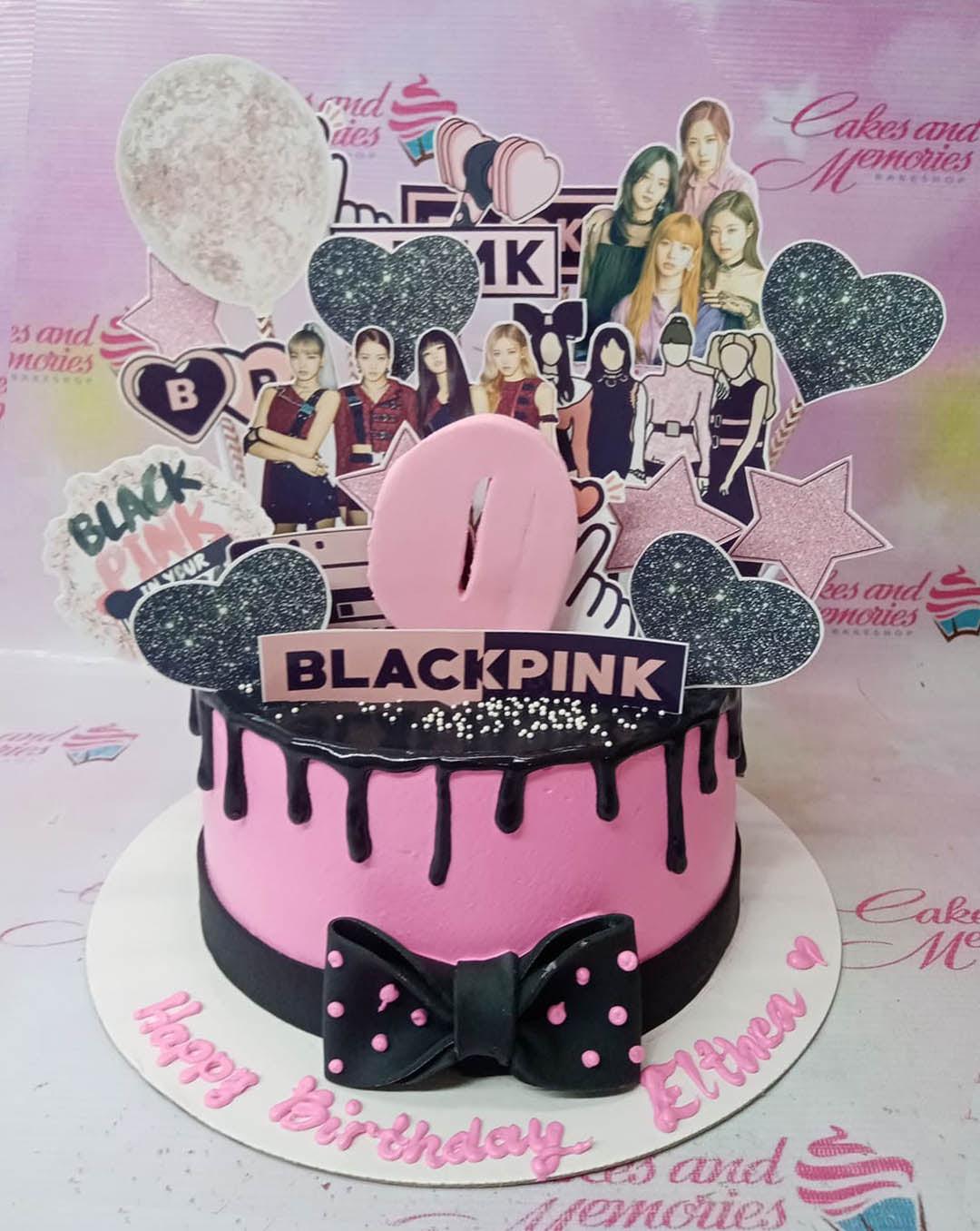 Pink Blackpink themed birthday cake with black chocolate drip, black bow, and various character printouts of the K-pop group members.