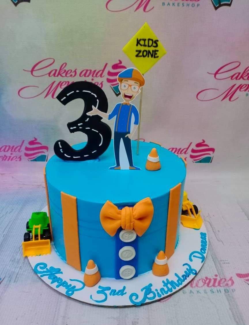Blue Blippi themed birthday cake with a large number 3 road topper, Blippi character cutout, and construction vehicle toys.