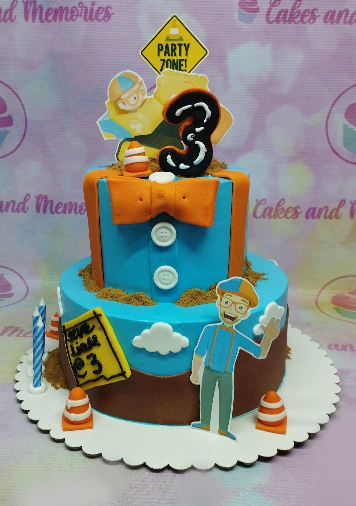 Two-tier blue and brown Blippi themed birthday cake with construction elements, orange bow, and character toppers.