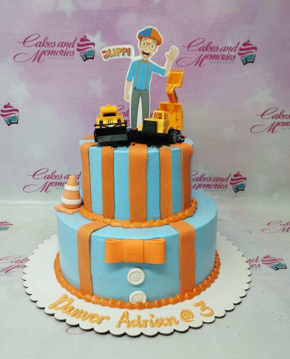 Two-tier light blue Blippi themed birthday cake with orange stripes, a fondant bow, and construction vehicle toppers.