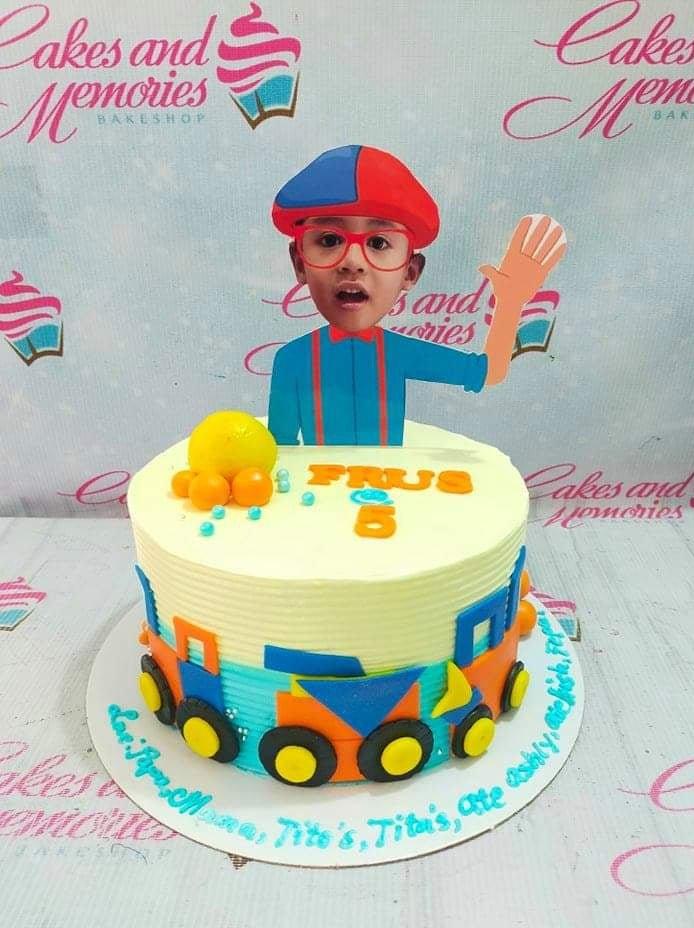 Blippi themed birthday cake with a custom photo printout topper and colorful fondant construction vehicles on yellow soft icing.