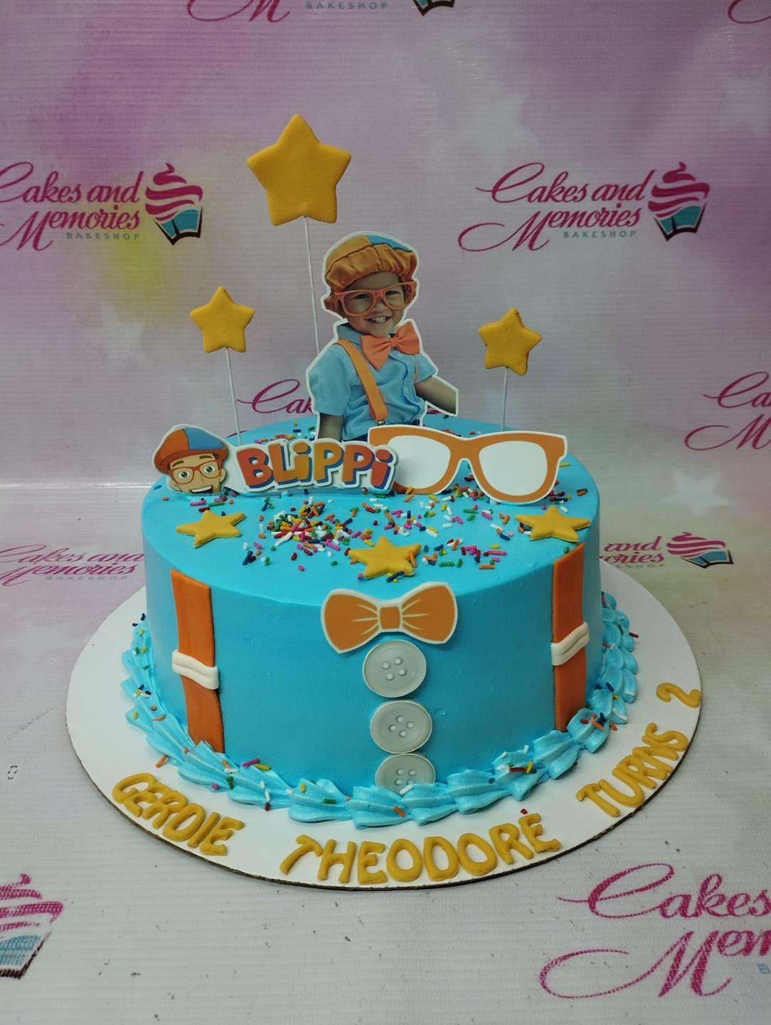 Blue Blippi themed birthday cake with orange suspenders, bow tie, and character toppers.