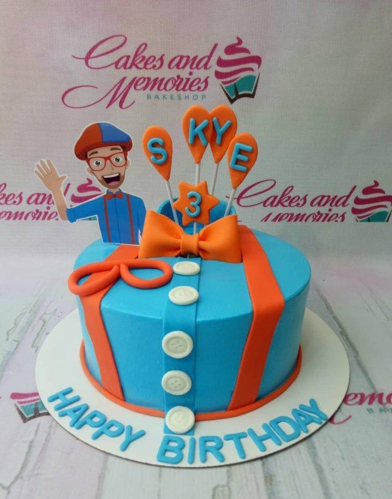 Blue Blippi themed birthday cake with orange suspenders, white buttons, and a Blippi character printout topper.