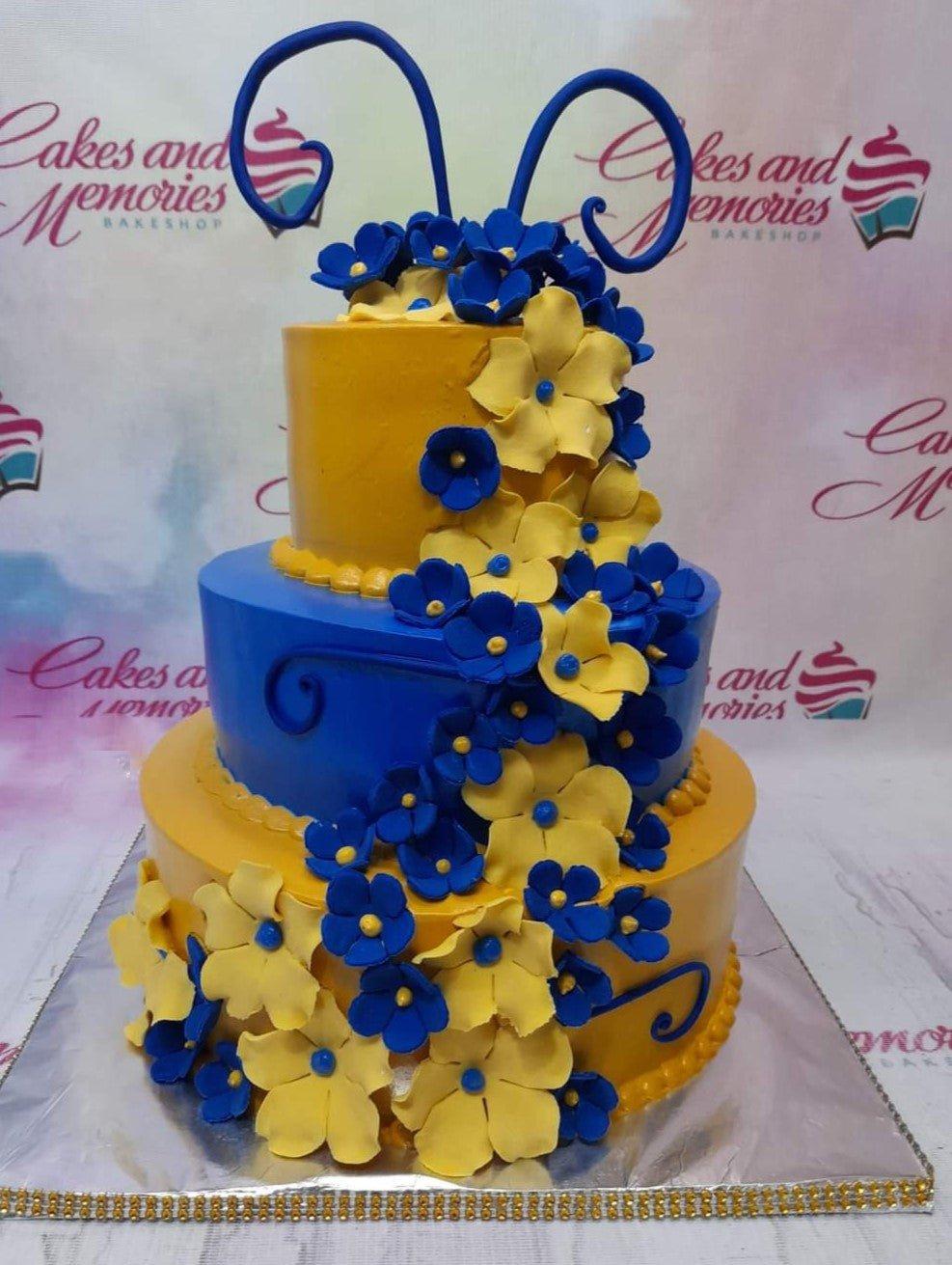 Three-tier blue and yellow floral cake with cascading gumpaste flowers and decorative blue swirls.