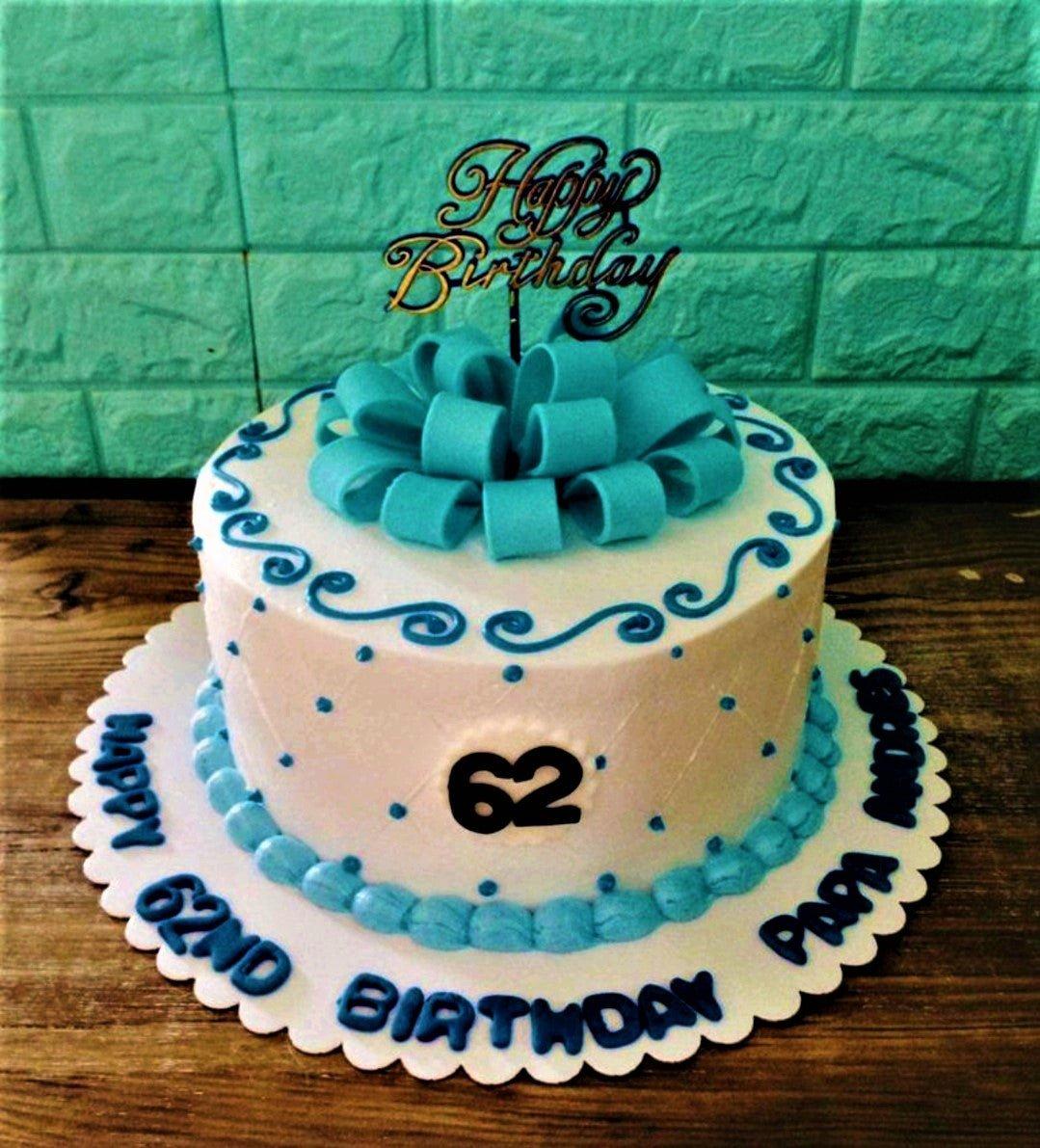 White 1-tier birthday cake with a large blue fondant bow, blue icing swirls, and a gold Happy Birthday topper.