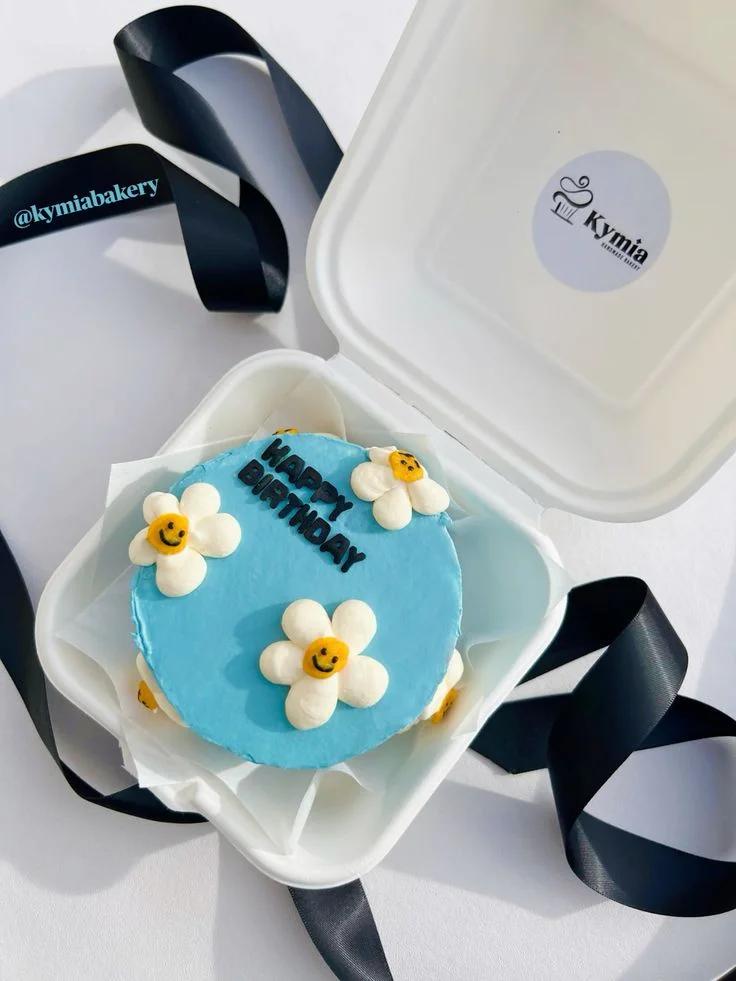 Light blue bento cake with white and yellow piped icing daisies and black Happy Birthday message in a white box.