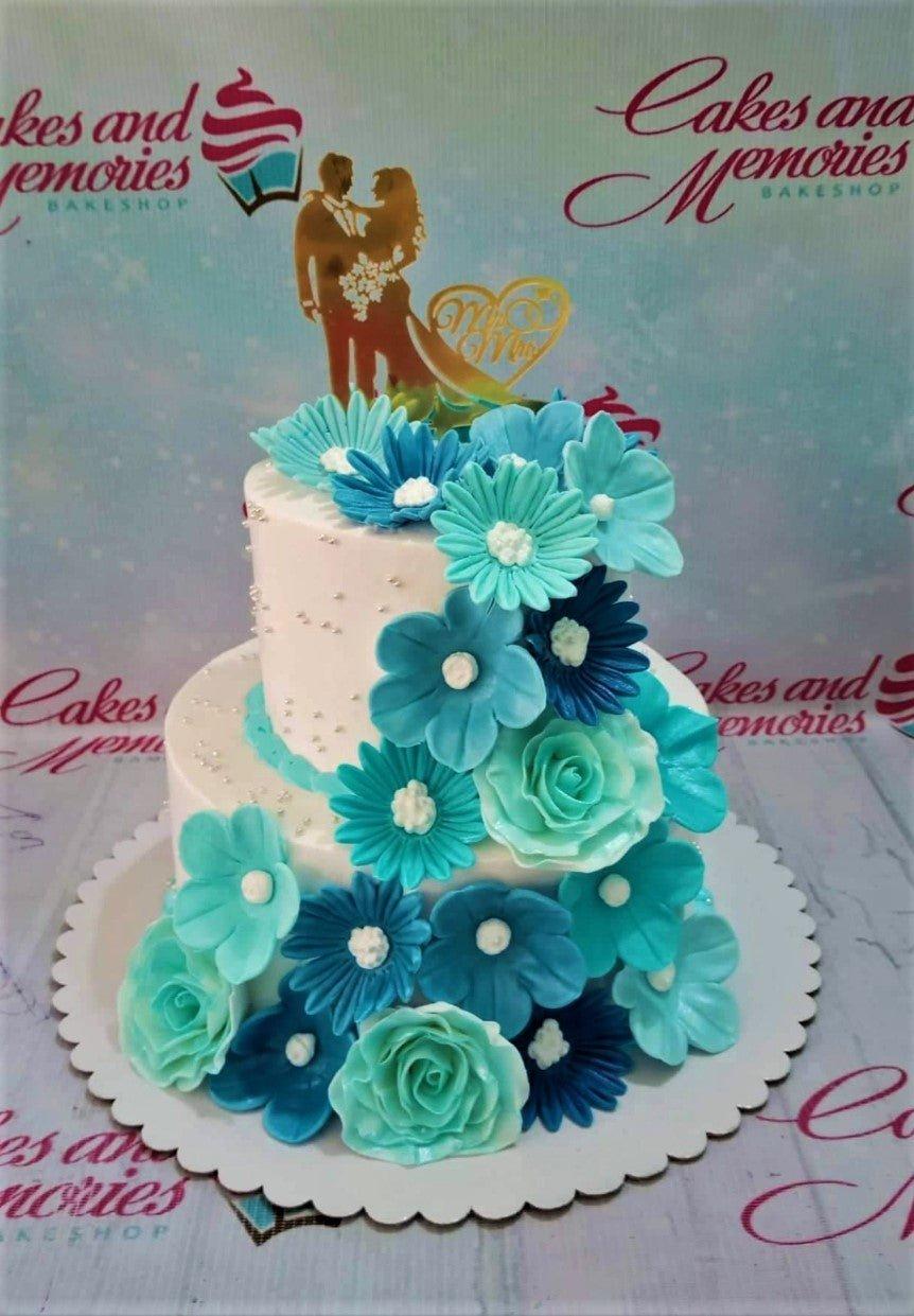 Two-tier white wedding cake decorated with a cascade of blue and teal gumpaste flowers and a gold couple topper.