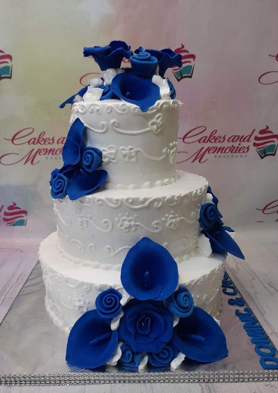 Elegant 3-tier white cake with intricate white icing swirls and vibrant blue gumpaste calla lilies and roses.