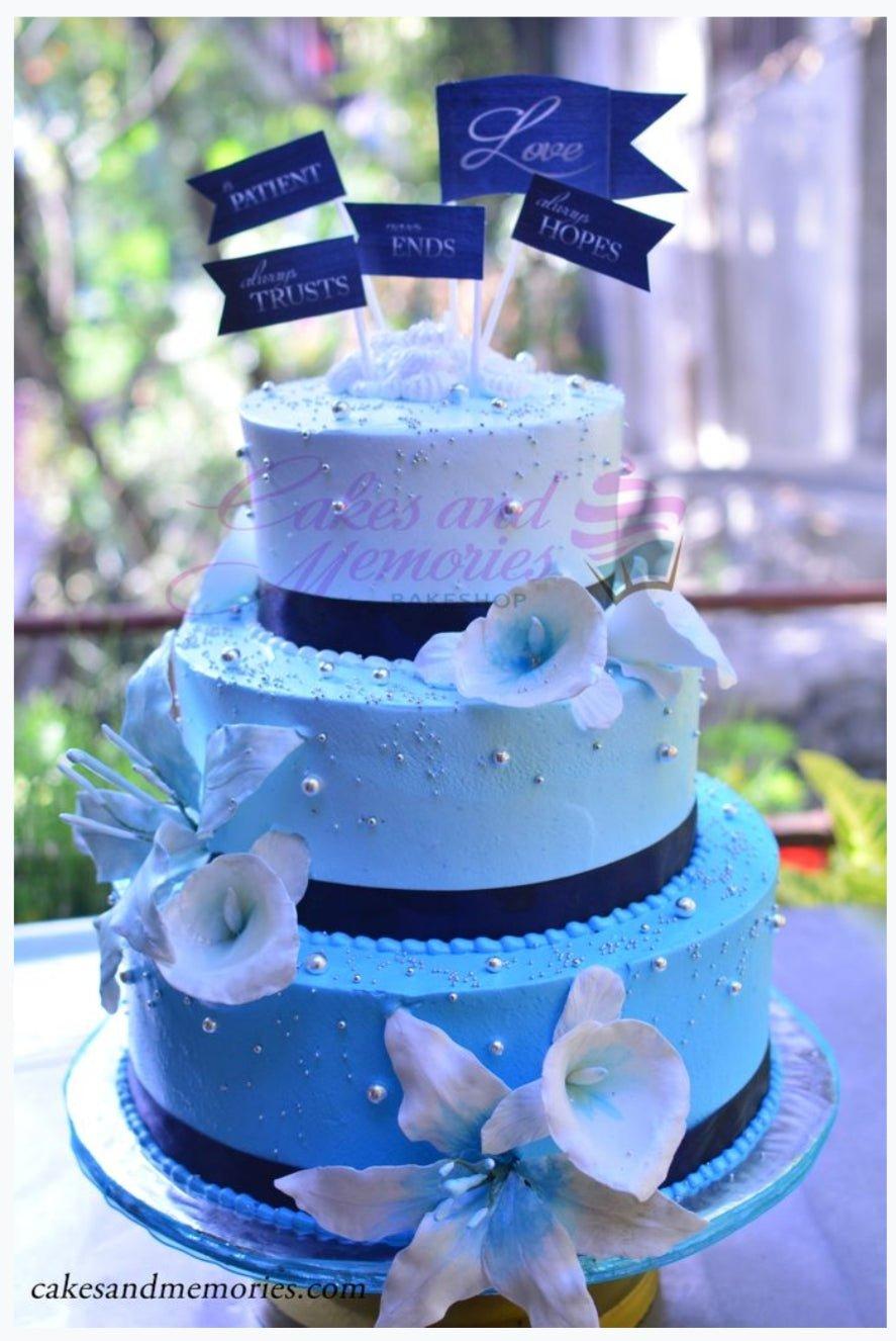 Three-tier blue gradient cake with white lilies, silver dragees, and blue flag toppers with inspirational messages.