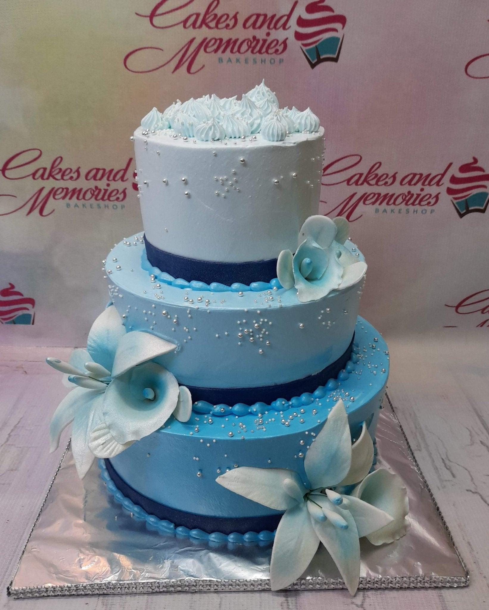 Elegant three-tier light blue ombre cake decorated with white and blue gumpaste lilies, silver dragees, and navy blue ribbon bands.
