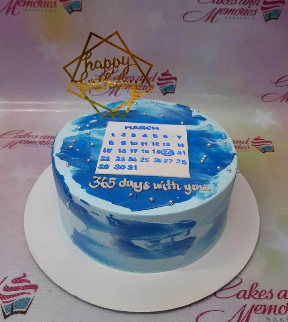 Light blue minimalist cake with dark blue palette knife icing strokes, a calendar printout, and a gold Happy Birthday topper.
