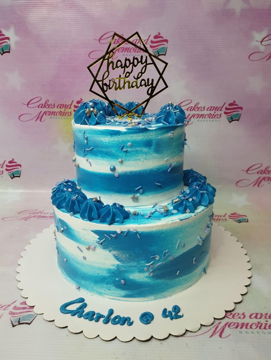 Two-tier blue and white watercolor cake with blue icing swirls, sprinkles, and a gold Happy Birthday topper.