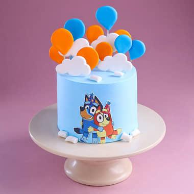 Light blue Bluey themed birthday cake with fondant balloons, white clouds, and a Bluey and Bingo character printout on the side.