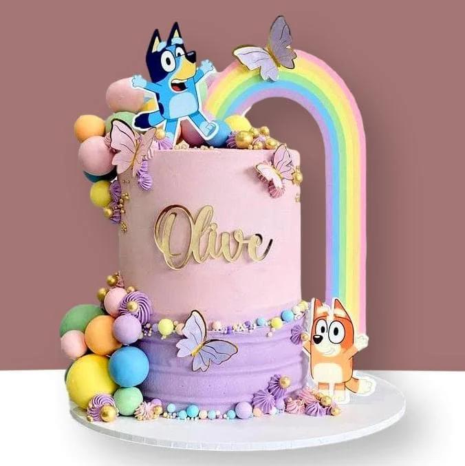 Purple and pink gradient cake with Bluey and Bingo characters, a rainbow backdrop, and colorful fondant spheres.