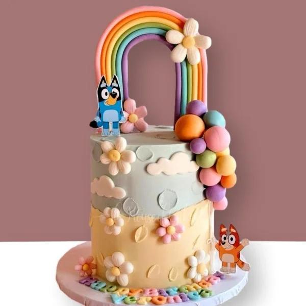 Bluey and Bingo themed birthday cake with a large rainbow topper, fondant flowers, and colorful balloon spheres.