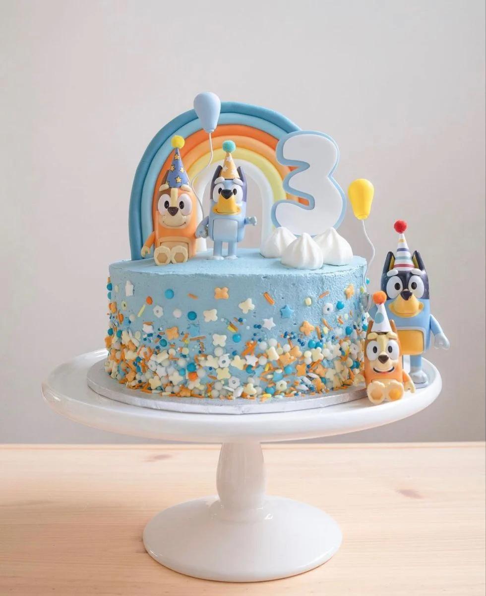 Bluey and Bingo themed light blue birthday cake with a fondant rainbow, white meringue clouds, and colorful sprinkles.