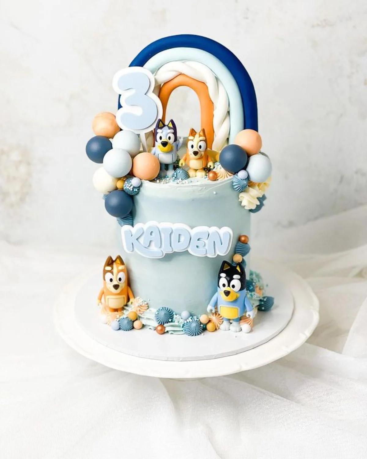 Light blue Bluey themed birthday cake with a fondant rainbow, 3D character figures, and colorful balloon toppers.