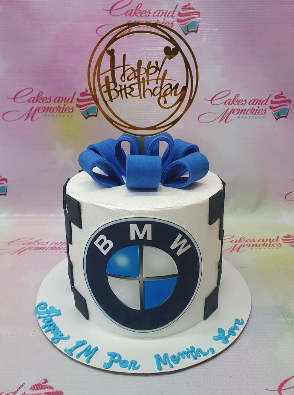 White 1-tier BMW themed birthday cake with a large blue fondant bow and a gold Happy Birthday topper.
