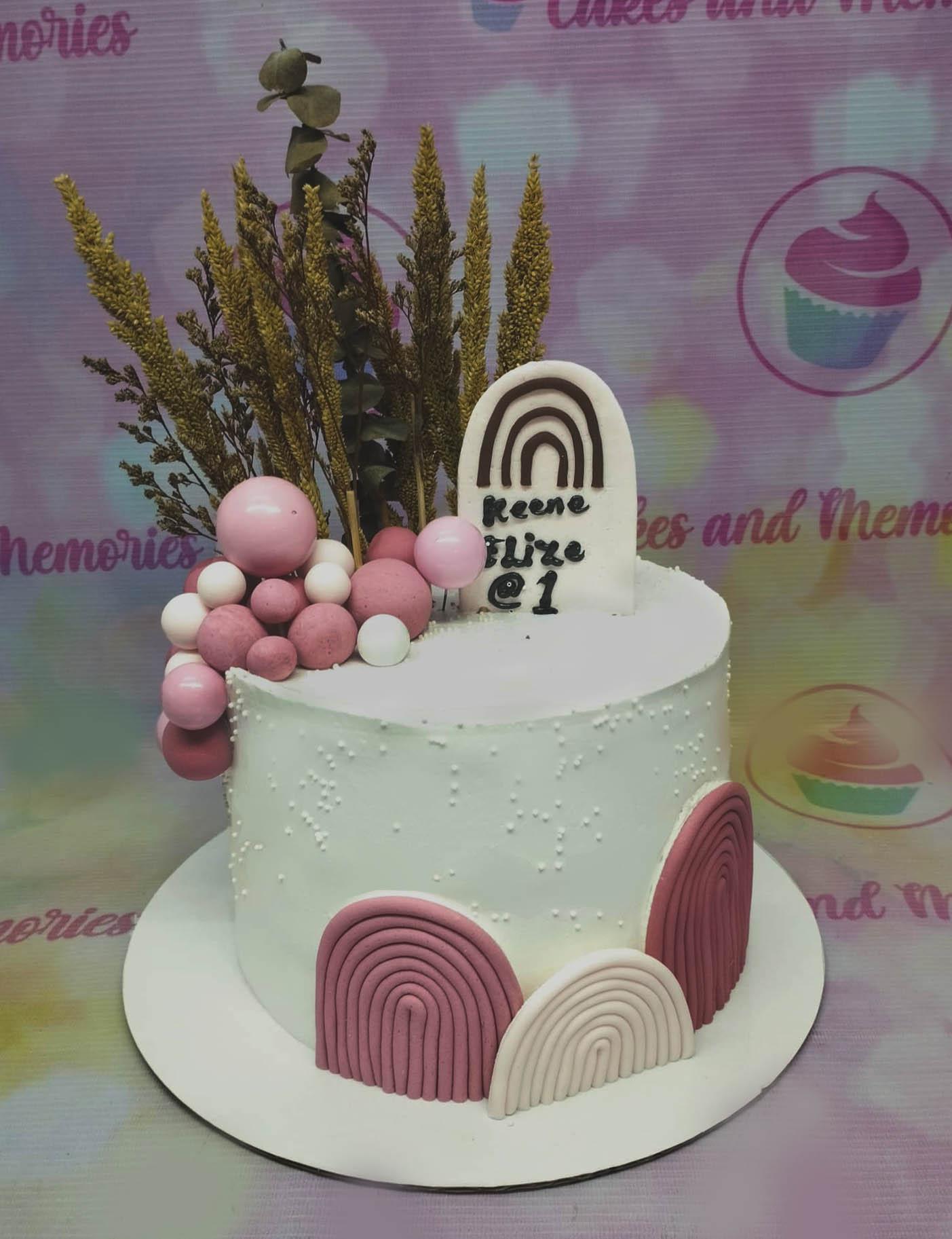 White minimalist cake with pink and white fondant balls, dried wheat stalks, and a boho rainbow topper for a first birthday.