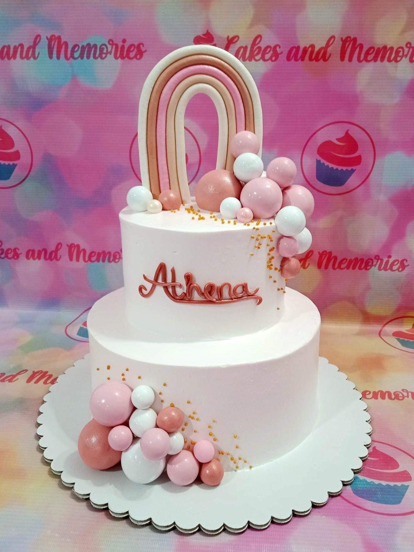 Two-tier white soft-icing cake with a large fondant rainbow topper, pink and white decorative spheres, and gold sprinkles.