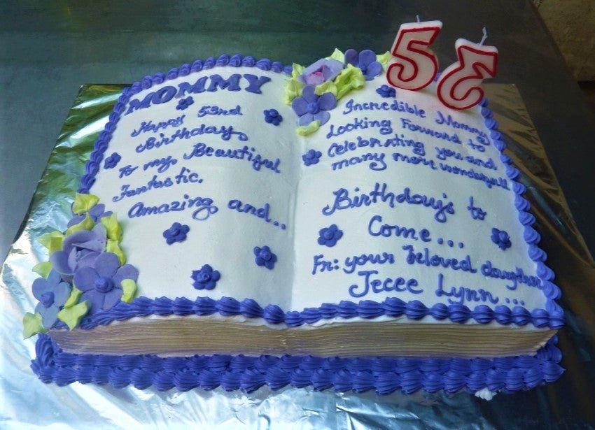 Open book shaped birthday cake with white icing, purple piped borders, and purple floral decorations for a 53rd birthday.