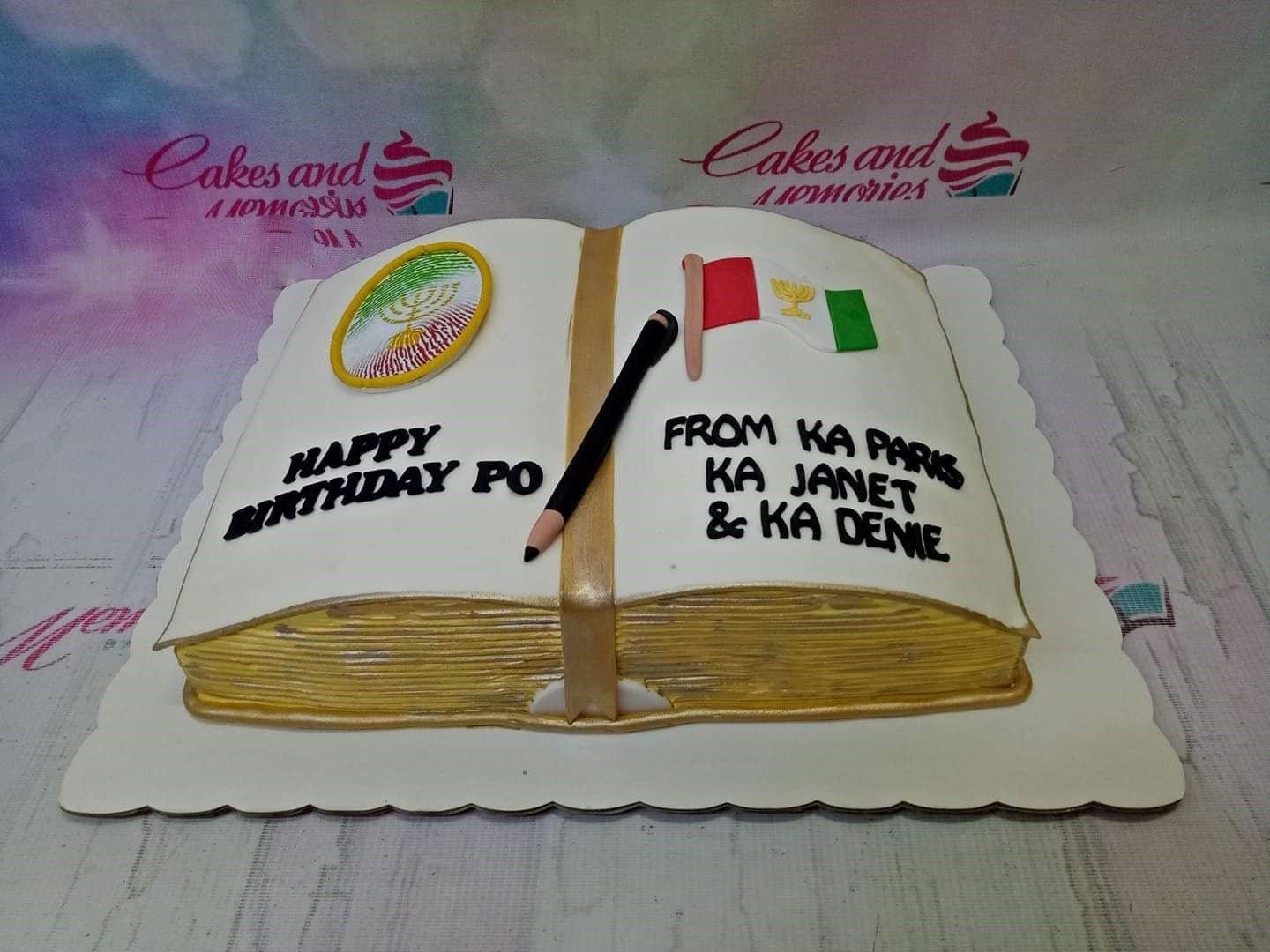 Open book shaped cake with white icing, gold pages, a pencil topper, and religious symbols for a birthday celebration.
