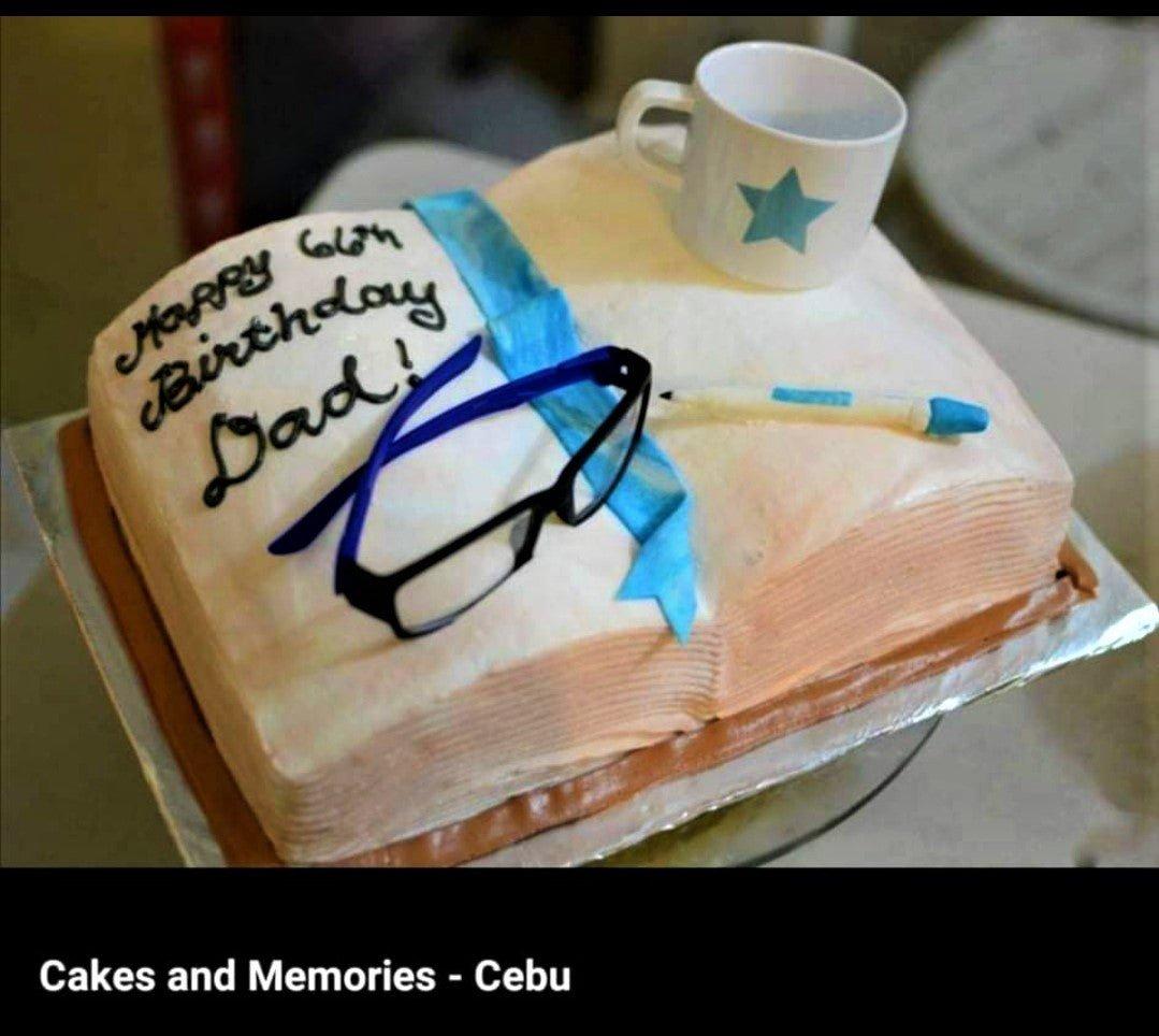 Square book-shaped cake for a 66th birthday with fondant glasses, a pen, a blue ribbon, and a white mug topper.