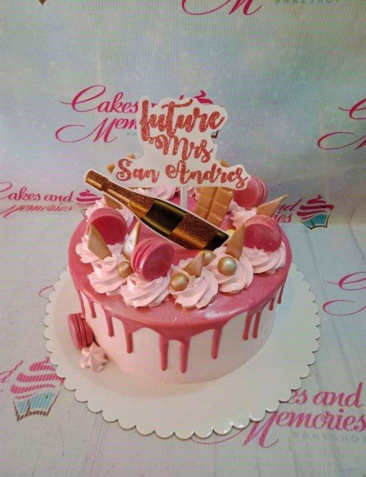 Pink 1-tier bridal shower cake with pink drip, macarons, chocolate bars, and a champagne bottle topper.