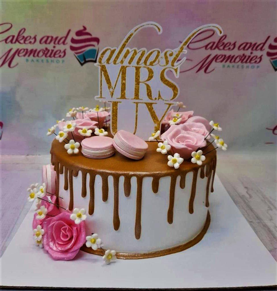 White 1-tier cake with gold drip, pink fondant roses, white daisies, pink macarons, and a gold glitter 'Almost Mrs Uy' topper.