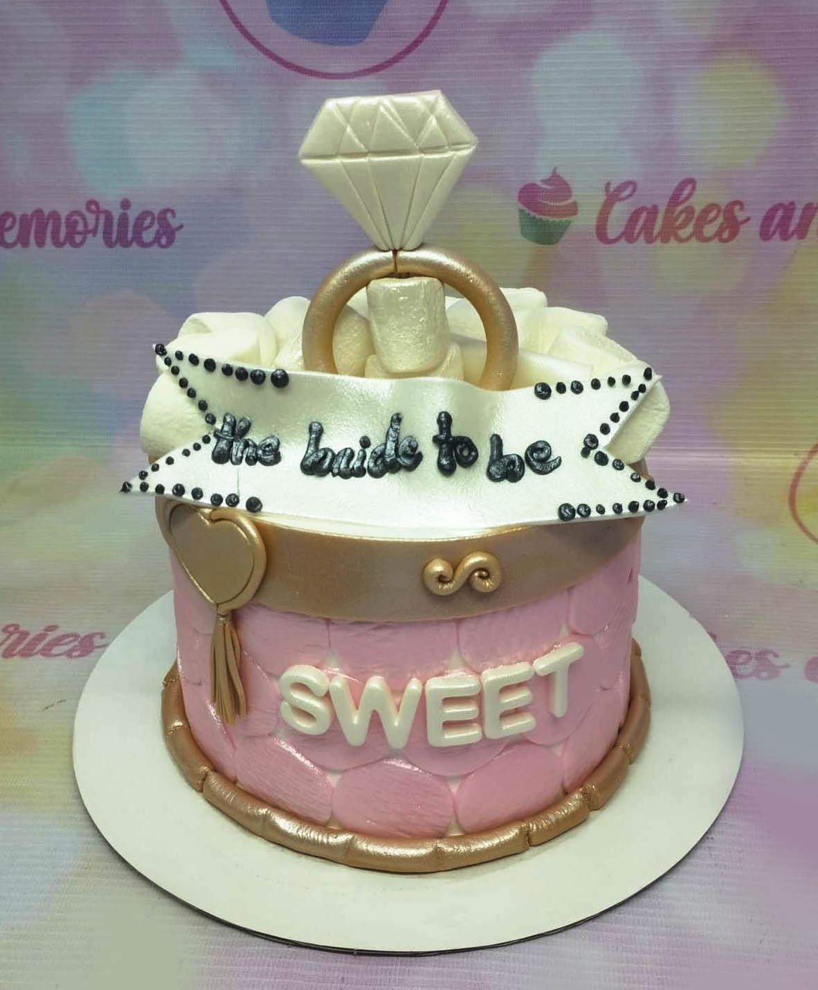 Pink bridal shower cake with a large diamond ring topper, white fondant banner, and gold accents.