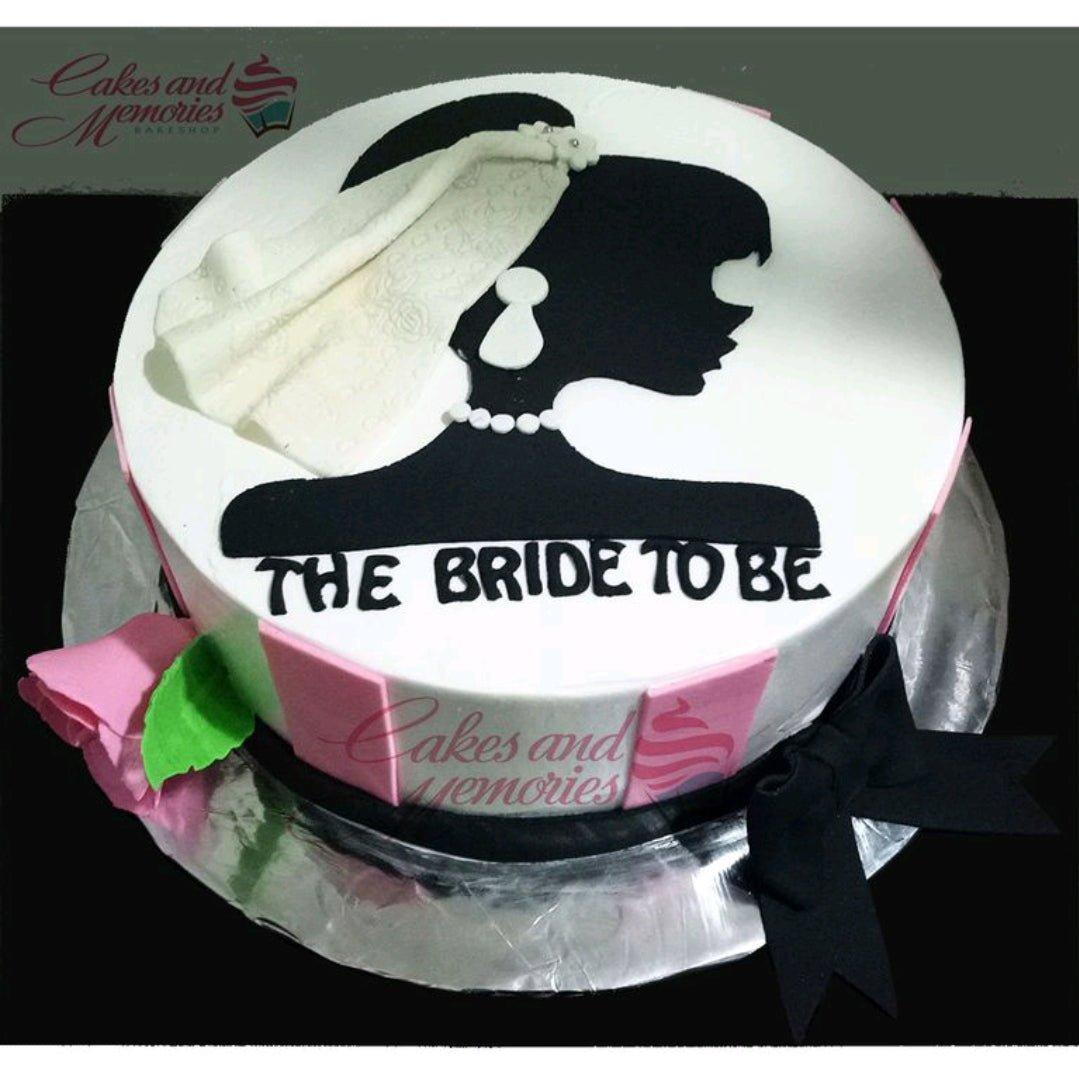 White round cake with a black silhouette of a bride wearing a white textured veil and pearl necklace, accented with pink side panels and a black bow.