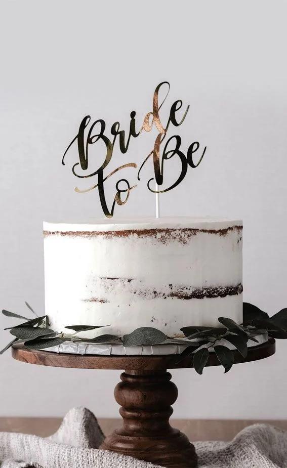 Semi-naked rustic white cake with a gold Bride to Be topper and green leaf accents on a wooden stand.