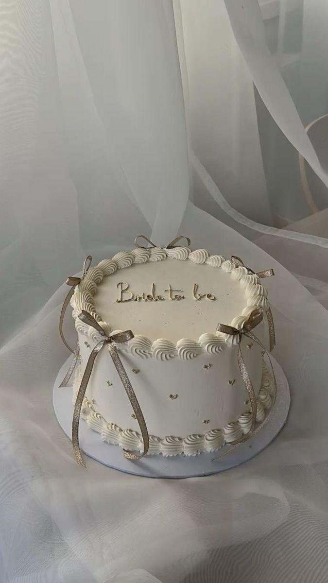 Elegant white minimalist bride-to-be cake with gold glitter bows and piped icing borders.