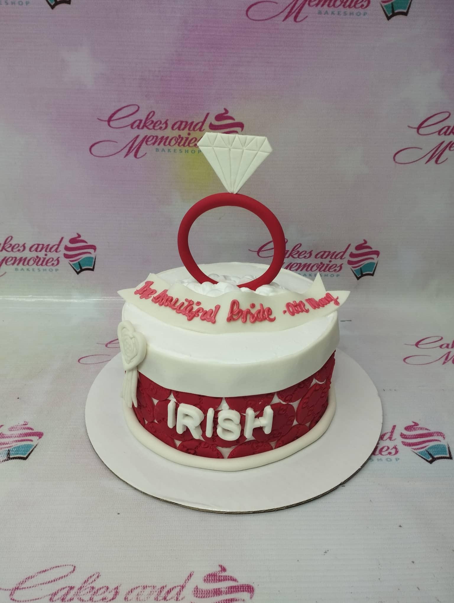 White and red fondant cake for a bride-to-be featuring a large diamond ring topper and a banner message.