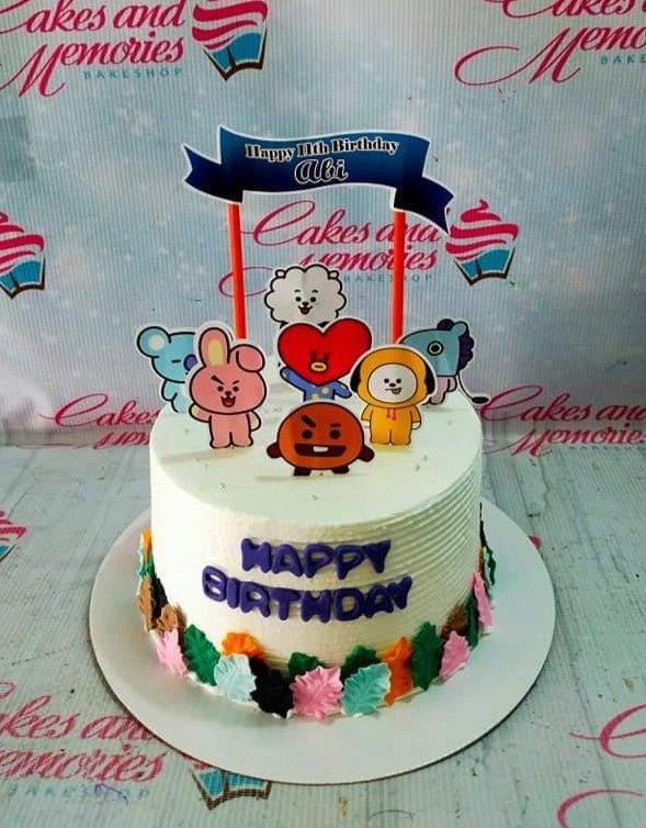 White soft-icing cake with BT21 character printouts, a blue birthday banner topper, and colorful icing leaves at the base.
