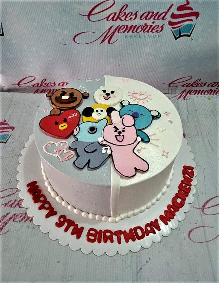 A round birthday cake with a split white and light blue icing base featuring BT21 character toppers and a message on the board.