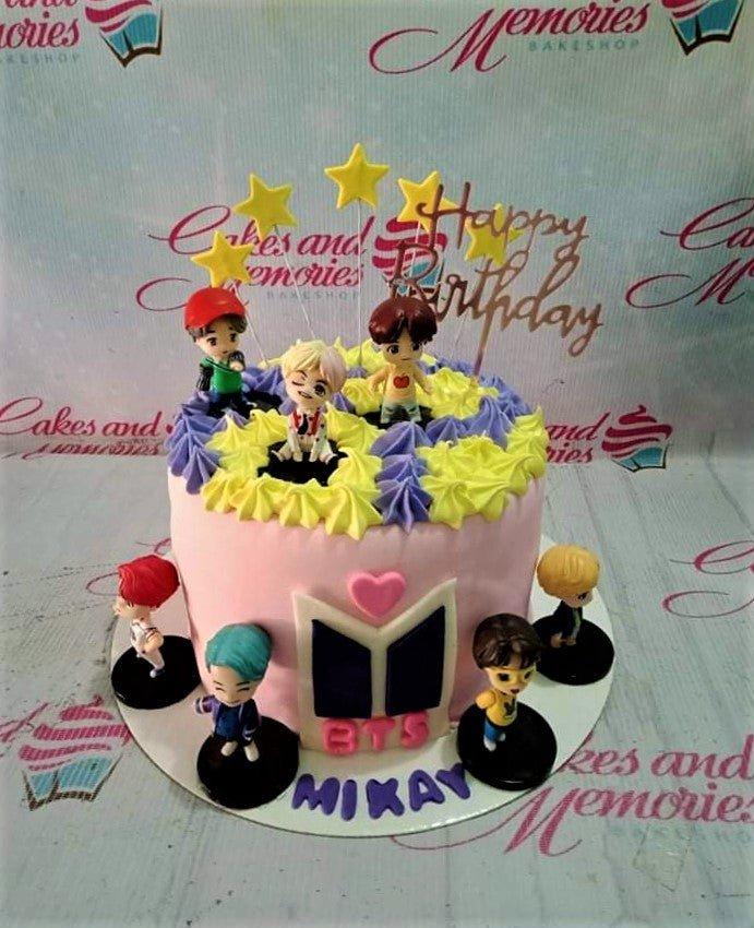 Pink BTS K-pop themed birthday cake with seven character figurines, yellow and purple icing rosettes, and gold star toppers.