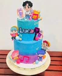 Two-tier blue and purple K-pop themed birthday cake with cartoon character printouts and musical note decorations.
