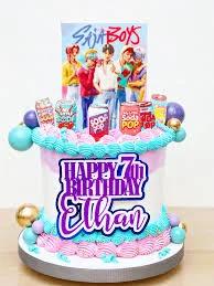Tall white and pink cake with blue icing borders, featuring K-pop boy band printouts and soda can toppers for Ethan's 7th birthday.