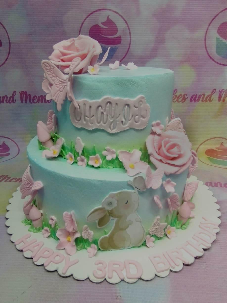 Two-tier light blue and green garden themed cake with pink roses, butterflies, and a cute bunny printout.