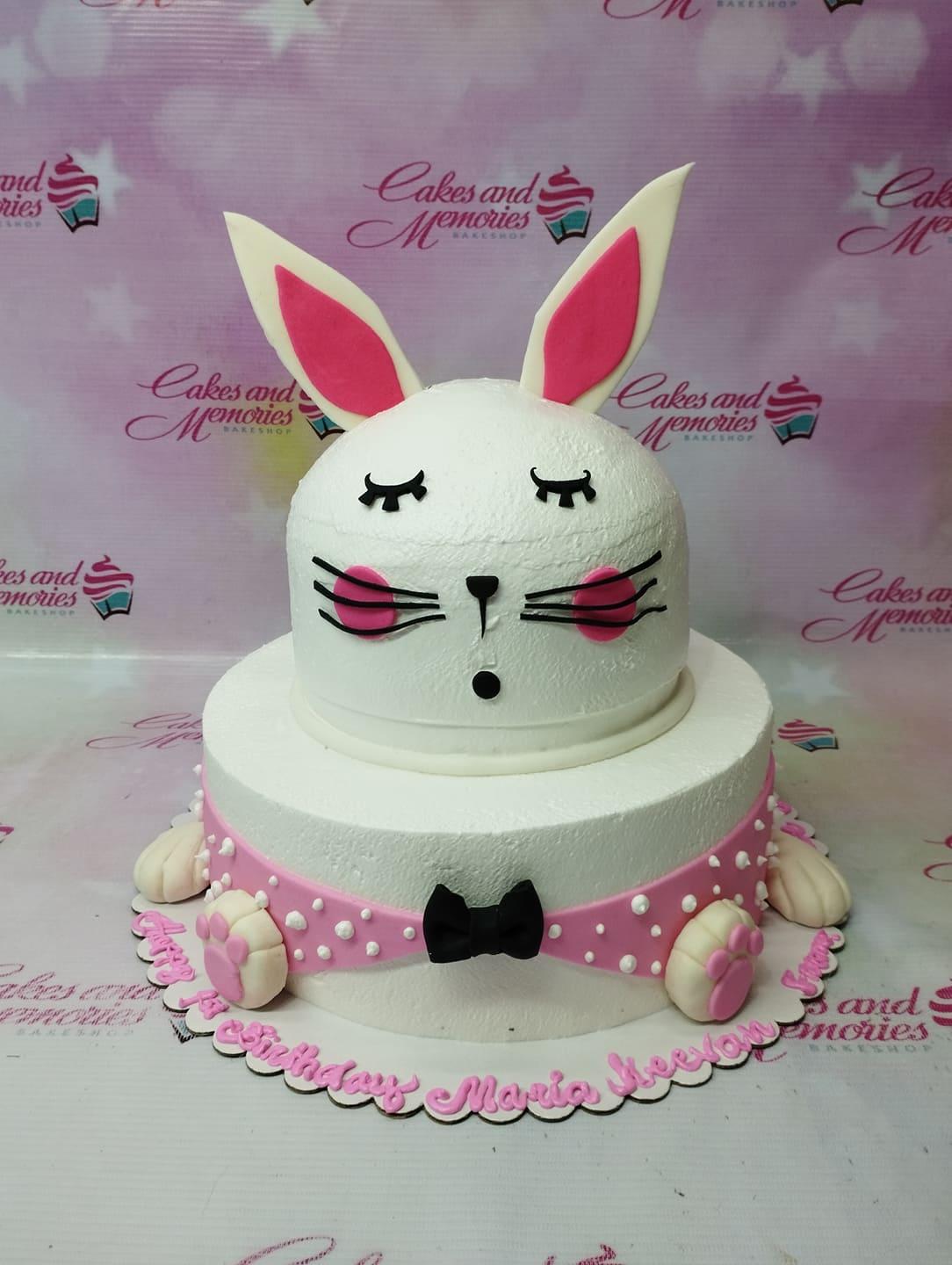 White 2-tier bunny-themed birthday cake with pink ears, black bow tie, and pink polka dot base wrap.