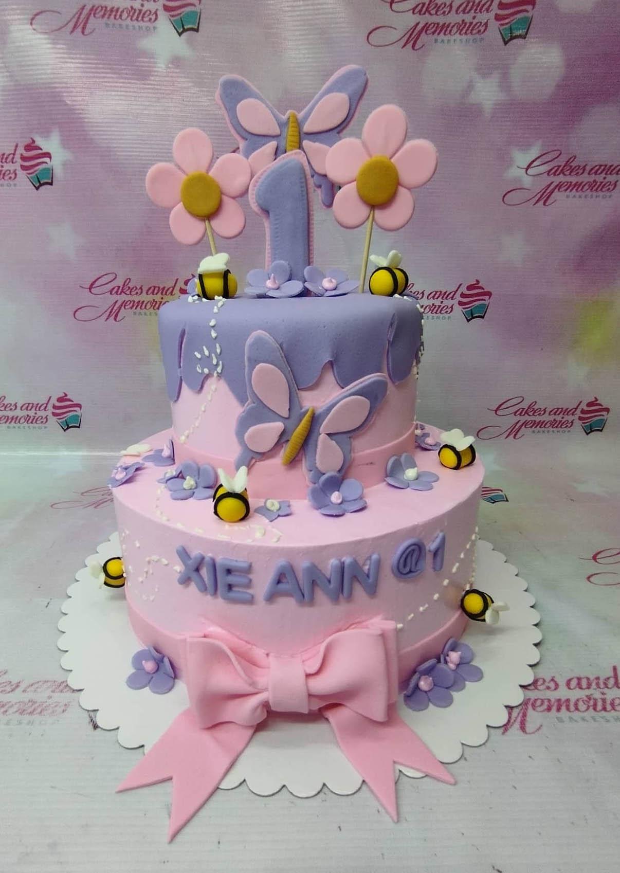 Two-tier pink and purple butterfly and bee themed birthday cake with fondant flowers and a large number one topper.