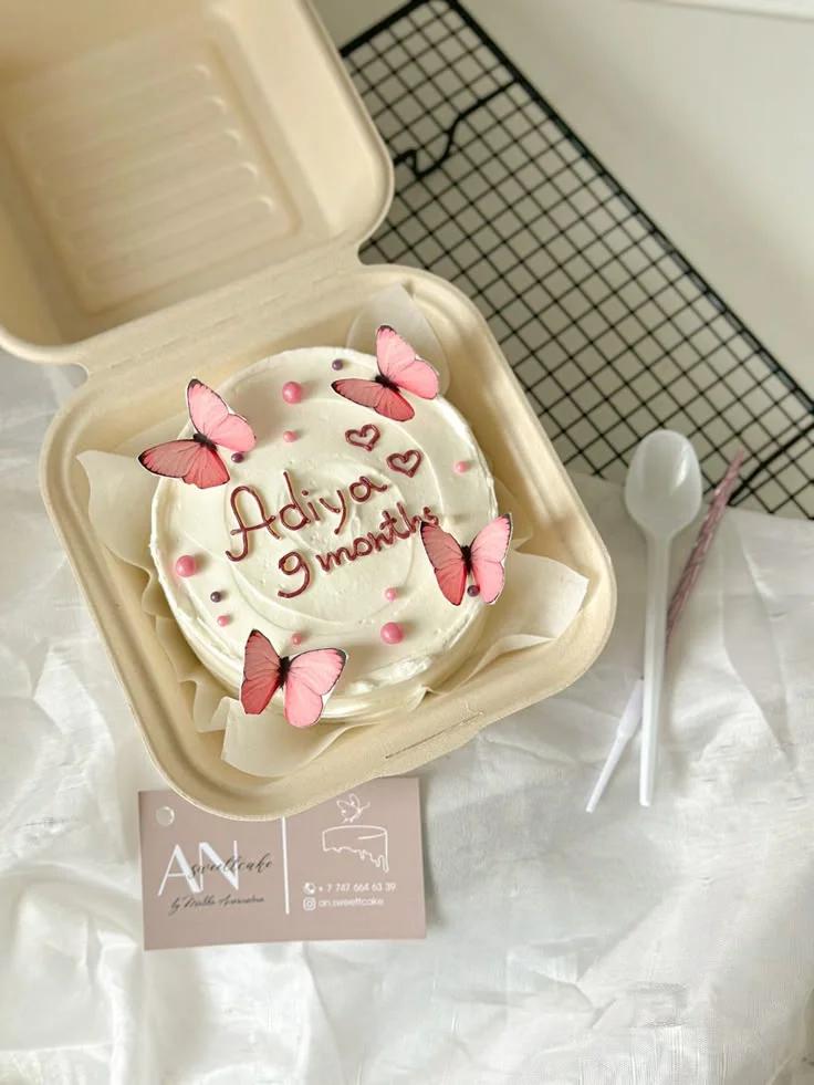 White bento cake in a box decorated with pink butterfly printouts, pink and purple pearls, and a message for a 9-month celebration.