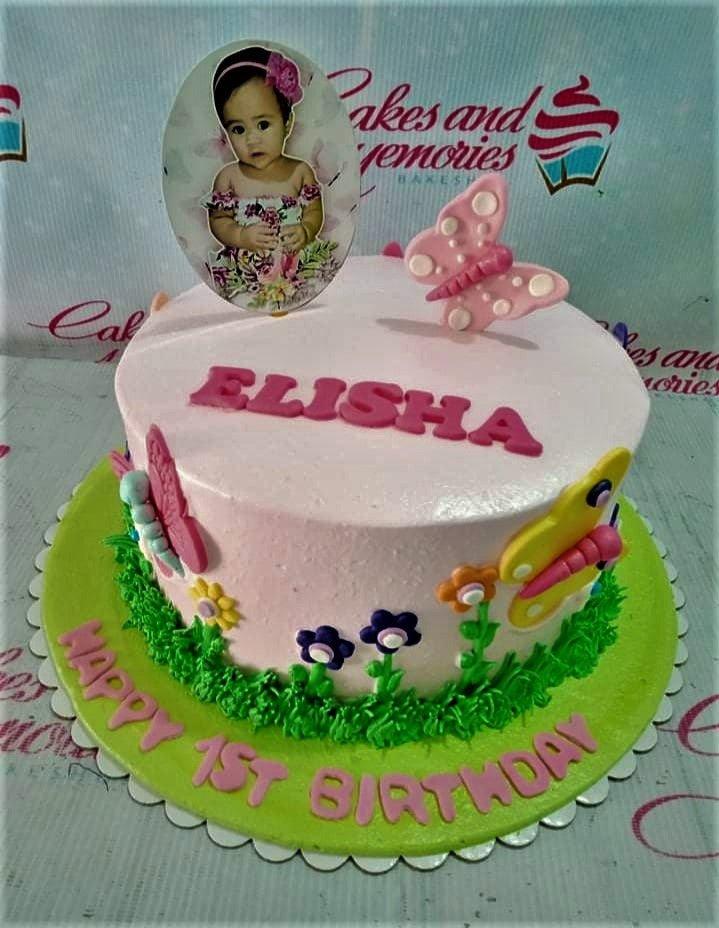 Pink 1st birthday cake for Elisha featuring a photo printout topper, pink butterfly, and green icing grass with colorful flowers.