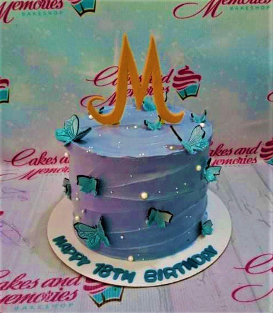 Purple and blue gradient cake with teal butterflies, white pearls, and a large gold letter M topper.