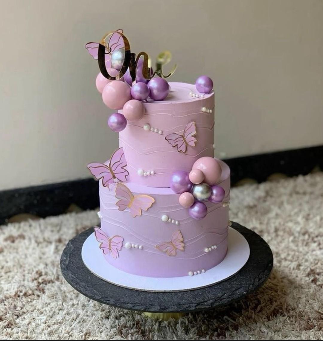 Two-tier lavender purple cake decorated with pink and purple butterflies, metallic spheres, and a gold 'One' topper.