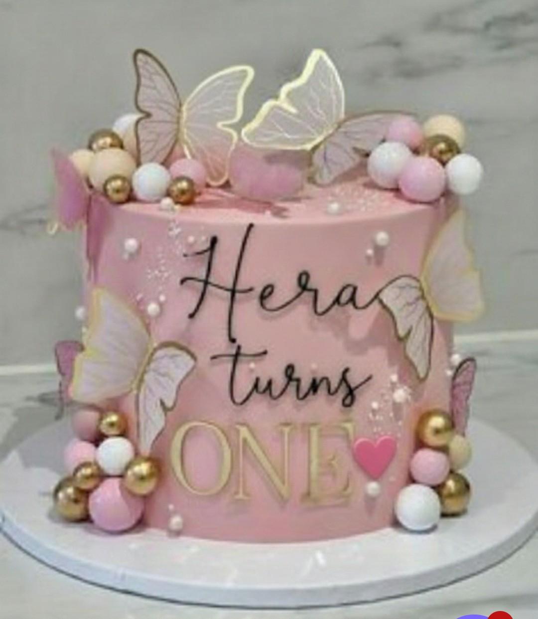 Pink butterfly birthday cake with gold and pink spheres and a personalized message for Hera's first birthday.