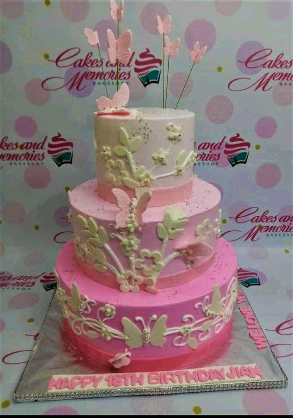 Three-tier pink gradient fondant cake decorated with white gumpaste flowers, leaves, and pink butterflies on wires.