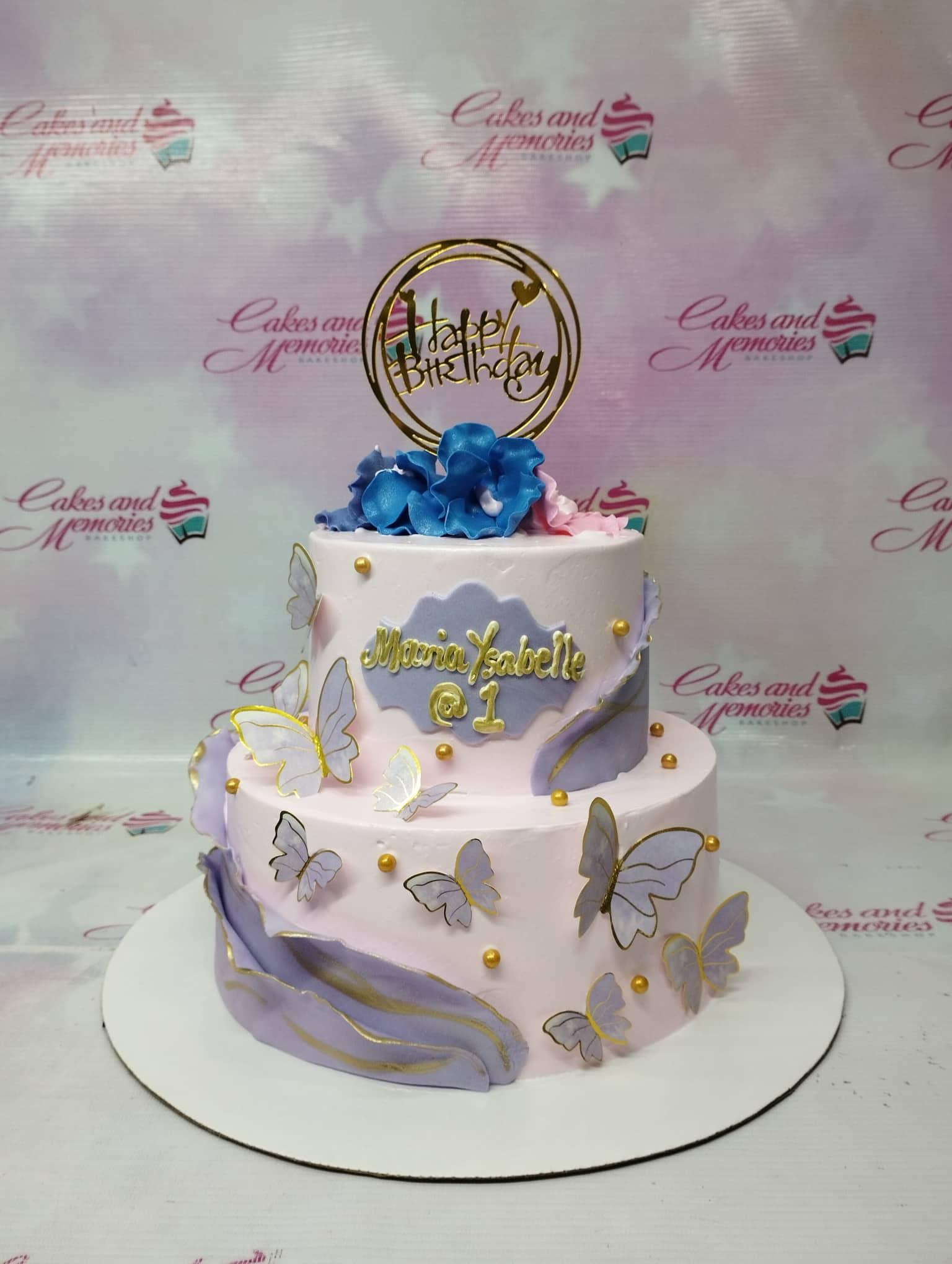 Two-tier light pink birthday cake with purple fondant drapes, gold butterflies, and a blue floral topper.