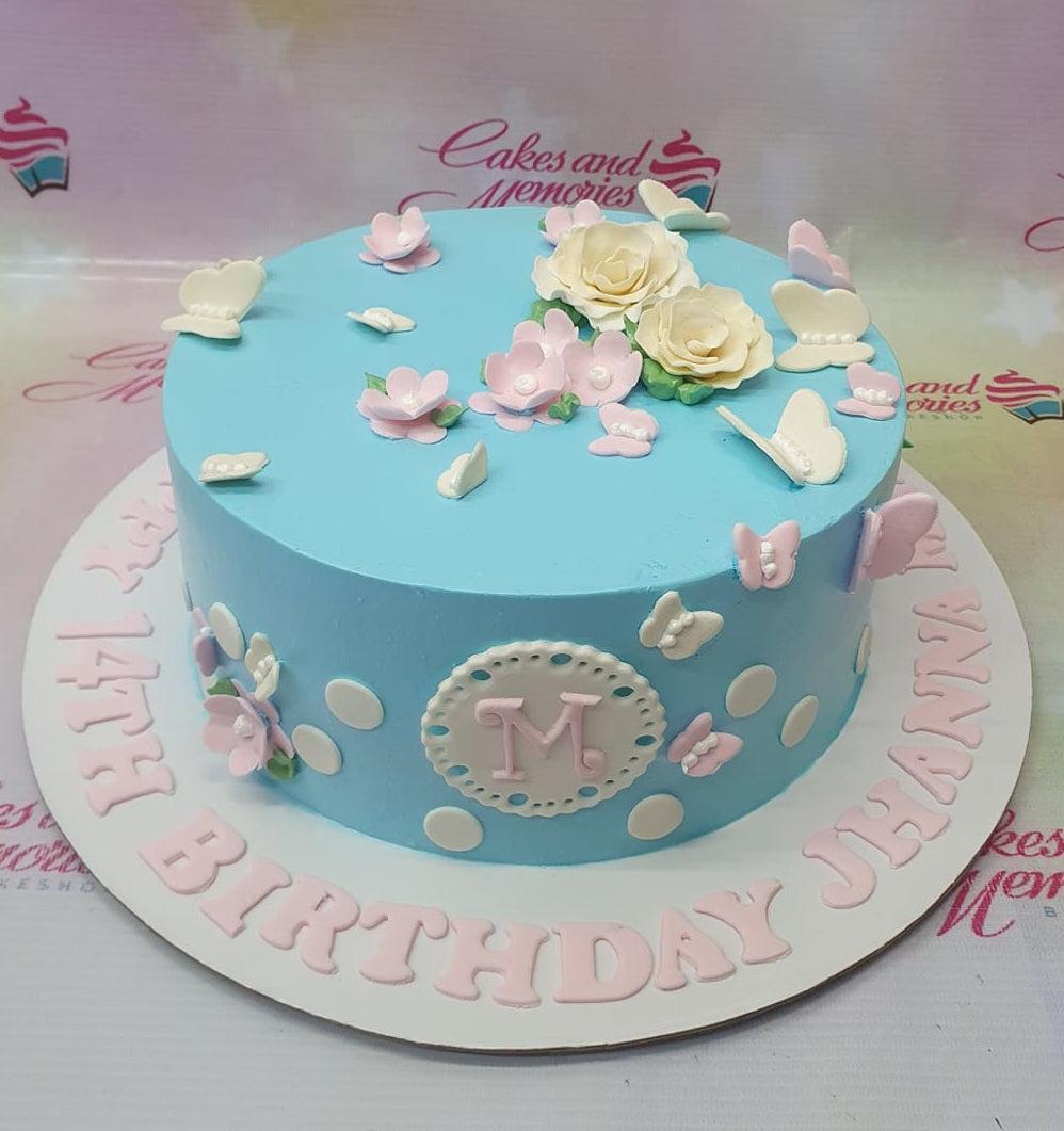 Light blue birthday cake with white and pink fondant flowers and butterflies, featuring a personalized 'M' monogram and birthday message.